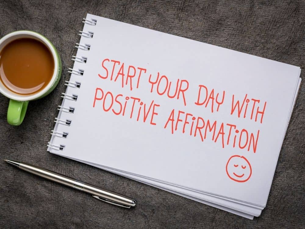 Notepad with Start your day with positive affirmation written in orange with a smiley face, lying on table next to silver pen and green coffee mug.